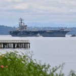A large aircraft carrier sailing on calm waters with a pier in the foreground and mountains in the distance.